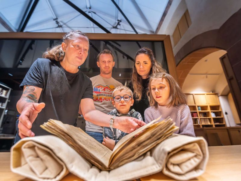 5 personnes (3 adultes, 2 enfants) regardent un grand livre ancien ouvert, protégé sur une table en bois. Une femme le tient. É#ères et vitrines en arrière-plan.